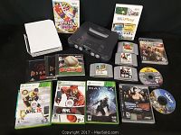 Photo shows Sega Genesis (black), Nintendo 64 (gray), and Wii (white) consoles alongside games for various platforms including Nintendo 64, Wii, Xbox 360, PlayStation 3, and PC.