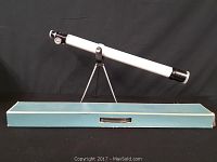 Side view of white refractor telescope mounted on slim chrome tripod stand with blue storage box underneath