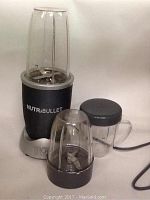 NutriBullet blender base with tall blending cup and blade assembly attached, shorter blending cup with lid beside it.