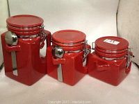 Photo showing three OGGI red glass kitchen canisters of varying sizes with metal lids, latches, and attached metal scoops.