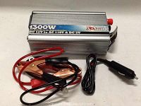 Photo shows XShade 1300 watt power inverter with aluminum casing, connected with clamp cables and cigarette lighter plug cable on a white surface.