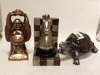 Photo of 3 decorative figures: bronze-colored Buddha, metallic horse head on checkered base, and dark turtle statue.