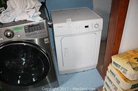Side view of the white Samsung dryer next to a washer and bags of cement, showing the door and partial control panel.