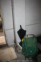 Fishing rods, a green backpack, black umbrella, and yellow hard hat grouped together near a wall.