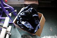 Dark air mattress folded inside cardboard box, showing vinyl surface and valve, placed on floor with exercise equipment in background.