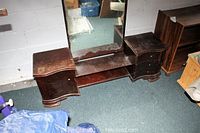 Photo of dark wood low boy dressing table with attached full-length mirror, showing worn surfaces and missing knobs, placed against white wall.