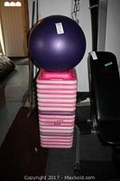 Stack of 18 small pink exercise steps with a large purple rubber exercise ball on top, next to an exercise bench.
