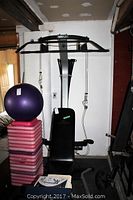 Full view of Bowflex Ultimate Home Gym with padded seat and backrest, overhead pull-up/dip bar, cable attachments, and pulleys. Visible purple exercise ball and pink aerobic steps stacked nearby.