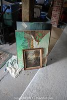 Three assorted framed prints and lithographs leaning against a wall, showing a waterfall scene and a smaller water and tree scene in front of it.