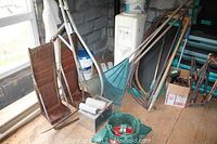 Image showing metal car ramps with rust, garden rakes, large spades and pruning saws leaning against a wall, with various other small garden tools on the floor.
