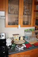 Kitchen counter and cabinets showing crystal wine glasses and glassware stored inside glass-front cabinet, with various kitchen and decorative items on counter.