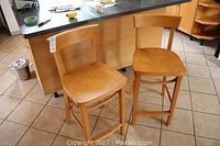 Pair of pine wooden bar stools from front and side views placed on tiled floor near kitchen island.