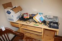 Photo of wooden desk with assorted electronics and stationery items piled on top including cables, chargers, remote controls, headphones, USB reader, envelopes, and CDs.