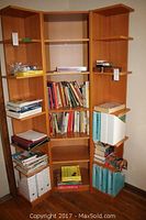 Wooden bookshelf with approximately 50 cookbooks, reference books, and notebooks arranged on shelves, some stacked on floor.
