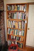 Full view of the wooden laminate bookcase filled with various books on all five shelves, standing next to a white door and a brick wall.