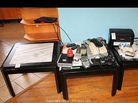 Wide view of two black tables showing many electronic devices including phones, remotes, printer, and clock radio arranged on top.