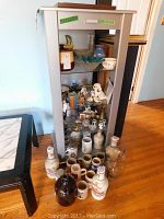 Photo of display cabinet containing various ceramic steins, dog figurines, bottles, and decorative glass bowl on hardwood floor