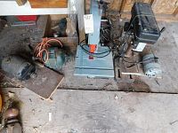 Photo showing Delta band saw, Rexon drill press, and three motors with visible rust and dirt on a wooden floor in workshop