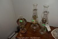Three clear glass oil lamps on wooden surface, sizes labeled as 13", 18", and 18.5".
