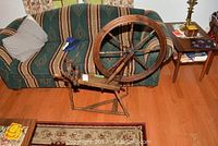 Full view of the antique wooden spinning wheel placed in front of a sofa on wooden floor, showing overall structure and size.