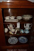 Shelves in wooden cabinet with various ceramic and glass kitchenware including bowls, teapots, plates, creamers, and cups