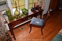 Wooden hall table with three drawers and brass hardware shown with decorative glassware on top. Blue upholstered wooden chair placed near the table.
