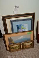 Five framed paintings grouped together on floor showing sizes and frames; largest watercolor by Alana Biasi visible with flowers and blue mat; mountain landscape painting and two smaller framed paintings also visible