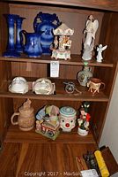 Full view of wooden shelf with various blue ceramic glassware, musical ornaments, cookie jars, angel figurines and small replica cannon arranged across three shelves.