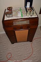 Vintage RCA Victor radio record player in wooden cabinet with tuning dial and speaker grille visible at front. Top surface has decorative items.