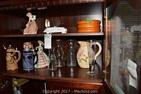 Wide shot showing all items inside glass-front cabinet including beer steins, figurines, milk bottle, wine carafes, and floral pitcher.