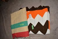 Two folded blankets side by side on carpet. Left is cream blanket with green and red stripes. Right is crochet afghan with zigzag pattern in brown, orange, white.