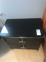 Black lacquer night stand with two drawers, silver trim and circular ring pull handles, standing on silver legs.