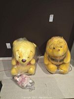 Pair of large yellow stuffed bears placed side by side on the floor, one with red collar and another with darker markings, both wrapped partly in plastic. Porcelain doll in pink dress lying in front, also in plastic.