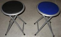 Photo showing two folding stools with round seats, one with a black seat and one with a blue seat, both on a concrete floor.