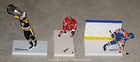 Photo shows three 6-inch hockey player figures: Mario Lemieux in black and yellow Pittsburgh Penguins uniform holding a trophy on a labeled white base; Eric Staal in red Carolina Hurricanes uniform skating on a small base; Wayne Gretzky in blue Edmonton Oilers uniform posed on a partial rink base with yellow and blue boards.