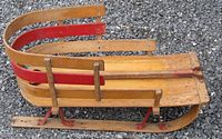 Side view of a wood and metal sled on gravel, showing curved wooden handle bars with red-painted metal parts and runners.