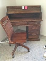 Wooden roll top desk with three drawers on right and center drawer, paired with a wooden office chair with cane back on casters, shown together.