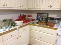 Overview of kitchen corner counter with green and clear glassware on left, red ceramic dishes and coffee/tea pots on right, against cream-colored cabinets
