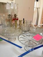 Display of two glass kerosene lamps, glass cake plates with patterns, bowls and brass candlesticks grouped together on a table.