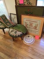 Full view of two small needlepoint stools, four framed textile artworks including a retirement-themed needlepoint and an embroidered pillow on the floor.