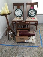 Photo showing two wooden chairs with needlepoint seats, two round clocks with bird and fish motifs, wooden plant stand, wooden magazine rack, and box with candles and candle accessories