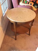 Round wooden table next to window and armchair, showing top and leg details with some wear marks.