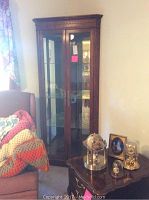 Wood corner cabinet with two glass doors and glass shelves inside. Decorative crown molding on top and brass handles. The cabinet is placed in a corner next to an armchair draped with crocheted blankets.