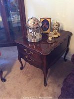 Brown Hammary wooden side table with two drawers. Decorative items on top including a rotating globe clock with a metallic base, a Bulova clock under a glass dome, a small pocket watch display, and a framed picture featuring dried plants arrangement.