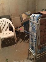 Photo showing a group of assorted chairs including plastic, metal, and woven styles next to a rollaway bed with floral mattress in a basement or storage area.