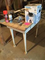 White wooden work table with worn paint and one drawer. The table holds various tools, a boxed kerosene lamp, a deadbolt lock, an antenna, and a shop flashlight with cord.