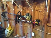 Two long-shaft gas-powered Weedeaters hanging on wall with visible engine sections and shoulder straps.