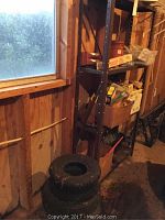 Metal shelving unit against wall with assorted hardware boxes and bags, visible work lights and circular saw blades on shelves, two tractor tires stacked on floor beside the shelf.