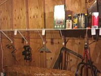 Tools hanging on wooden wall, showing hand saw, axe, hoe, sling blade, manual drill, pair of loppers and Coleman lantern along with cans of fuel and sprays on shelf.