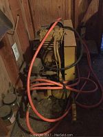 Air compressor unit placed on the floor against a wood wall with orange and black hoses coiled on top.
